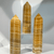 Three Orange Calcite towers standing upright, side-by-side. The polished mineral pieces have a six-sided, hexagonal shape and taper to a point, displaying distinct horizontal stripes of deep golden-yellow, warm orange, and white. Three Orange Calcite towers standing upright, side-by-side. The polished mineral pieces have a six-sided, hexagonal shape and taper to a point, displaying distinct horizontal stripes of deep golden-yellow, warm orange, and white.