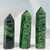 Three polished crystal towers, or points, made of Ruby Zoisite. The hexagonal towers are primarily bright green with heavy black flecks, characteristic of the mineral Zoisite. The crystals stand upright against a white background, varying slightly in height and width.