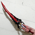 A close-up of a hand gripping the hilt of the Kamish's Wrath Dagger replica from Solo Leveling. The striking weapon has a vibrant red blade with black accents and a menacing black dragon-claw crossguard. The background is a white brick wall.