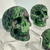 A profile view of a detailed skull hand-carved from green and pink Ruby Zoisite gemstone. The skull features a mottled green surface with distinct deep ruby-red spots and sits on a clear acrylic display stand.