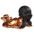 A small figurine of a smiling, young monk with a black finish, lying on his stomach in a relaxed pose. He is propping his chin up with both hands, showing a joyful expression with closed eyes and chubby cheeks. He wears a flowing, metallic copper-toned robe that drapes around his body, with his feet kicked up playfully behind him.