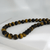 A high-detail, low-angle shot of a Tiger's Eye beaded necklace resting on a white surface. The 12mm round beads are highly polished, reflecting light and highlighting the natural fiber-like stripes and rich amber tones of the gemstones. The necklace curves across the frame, showcasing the consistent size and quality of the stones.