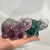 A pair of small, hand-sized elephant figurines carved from translucent fluorite gemstone. One elephant is a deep violet-purple, and the other is a rich forest-green, both featuring natural cloudy white and clear inclusions within the mineral. The elephants are sculpted with detailed ears, sturdy legs, and their trunks raised high in the air, catching the light to showcase the glossy, polished texture of the crystal.