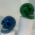 A collection of miniature decorative skulls displayed on white risers. Each high-gloss skull features a solid, opaque finish in vivid tones—including deep cobalt blue and emerald green—surfaced with striking, branch-like vein patterns that mimic cracks of lightning. The stylized skulls feature deep, dark eye sockets and realistic teeth detailed in crisp white and silver.