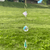 Hanging gold-tone sun catcher with three diamond-shaped frames, lavender beads, and faceted iridescent crystals against a blurred green lawn.