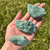 Hand holding three rough Fuchsite mineral specimens, displaying their natural green color, irregular shapes, and distinctive textured surfaces.