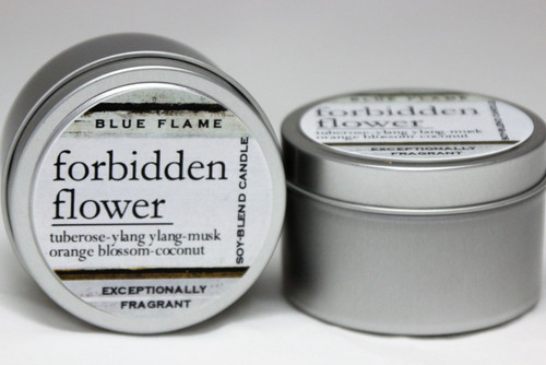 There are two silver, modern designed travel tins in the image. One tin is on it's side to show what the top of the label looks like on the lid: It says, forbidden flower underlined in the middle of the label, and is off set towards the left side in large text, tuberose-ylang ylang-musk-orange blossom-coconut are written below in a smaller font. The Blue Flame logo is at the top of the label. Soy-blend Candle is written on the right side in a vertical line. On the bottom of the label is written, Exceptionally Fragrant. The other tin is flat and shows that the tin is just silver without a wrap around label. The modern design uses pale hues and simplistic lines.