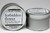 There are two silver, modern designed travel tins in the image. One tin is on it's side to show what the top of the label looks like on the lid: It says, forbidden flower underlined in the middle of the label, and is off set towards the left side in large text, tuberose-ylang ylang-musk-orange blossom-coconut are written below in a smaller font. The Blue Flame logo is at the top of the label. Soy-blend Candle is written on the right side in a vertical line. On the bottom of the label is written, Exceptionally Fragrant. The other tin is flat and shows that the tin is just silver without a wrap around label. The modern design uses pale hues and simplistic lines.