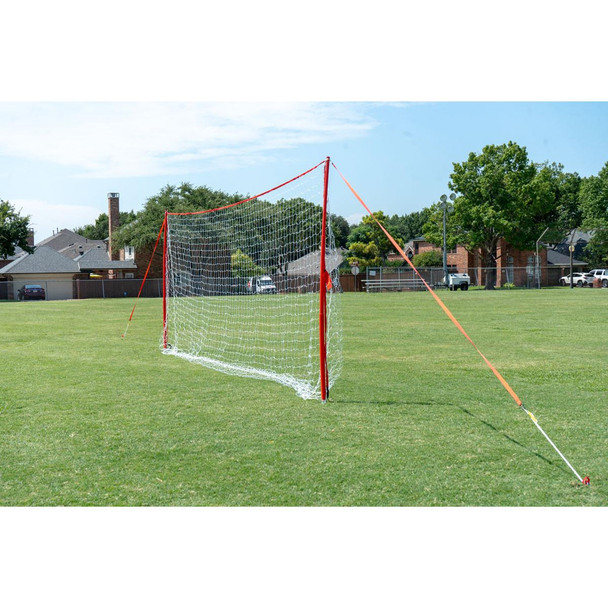 Regulation-size J-Goal portable soccer training goal with red frame, white net, and guy cords for stability, set up on a grassy field in a suburban park.