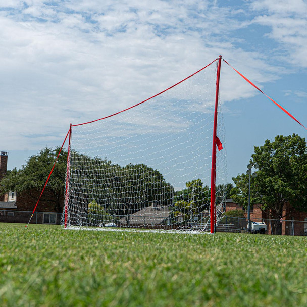 Regulation-size J-Goal portable soccer training goal with red frame and net, set up on a grassy field under a partly cloudy sky.
