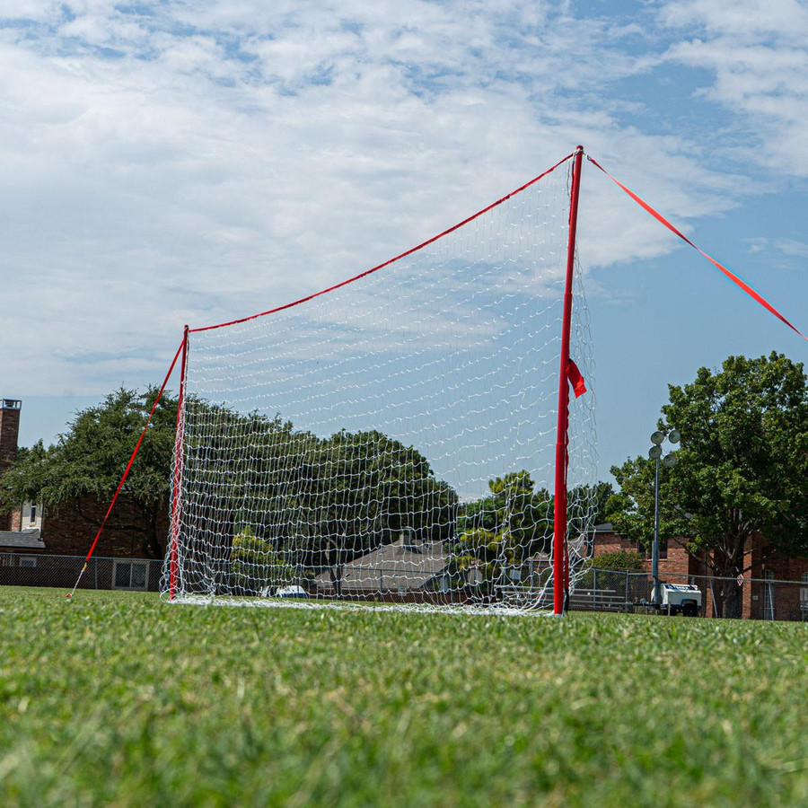 Regulation-size J-Goal portable soccer training goal with red frame and net, set up on a grassy field under a partly cloudy sky.