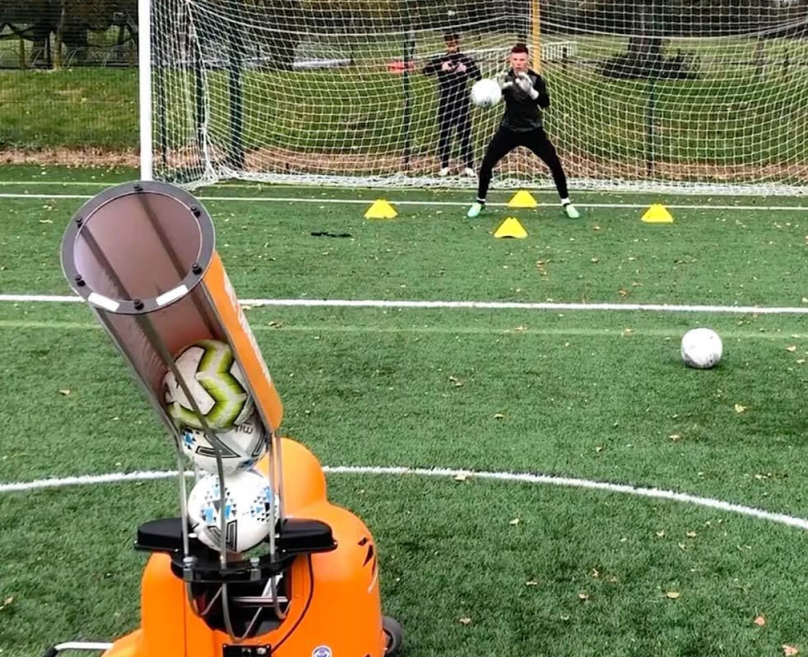 Automatic soccer ball launcher aimed at a goalkeeper in training on an outdoor turf field