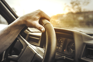 A black, 3” rigid seatbelt extension is buckled in to a seat belt on both ends, securing a man wearing jeans and a red shirt in the driver's seat of his car.