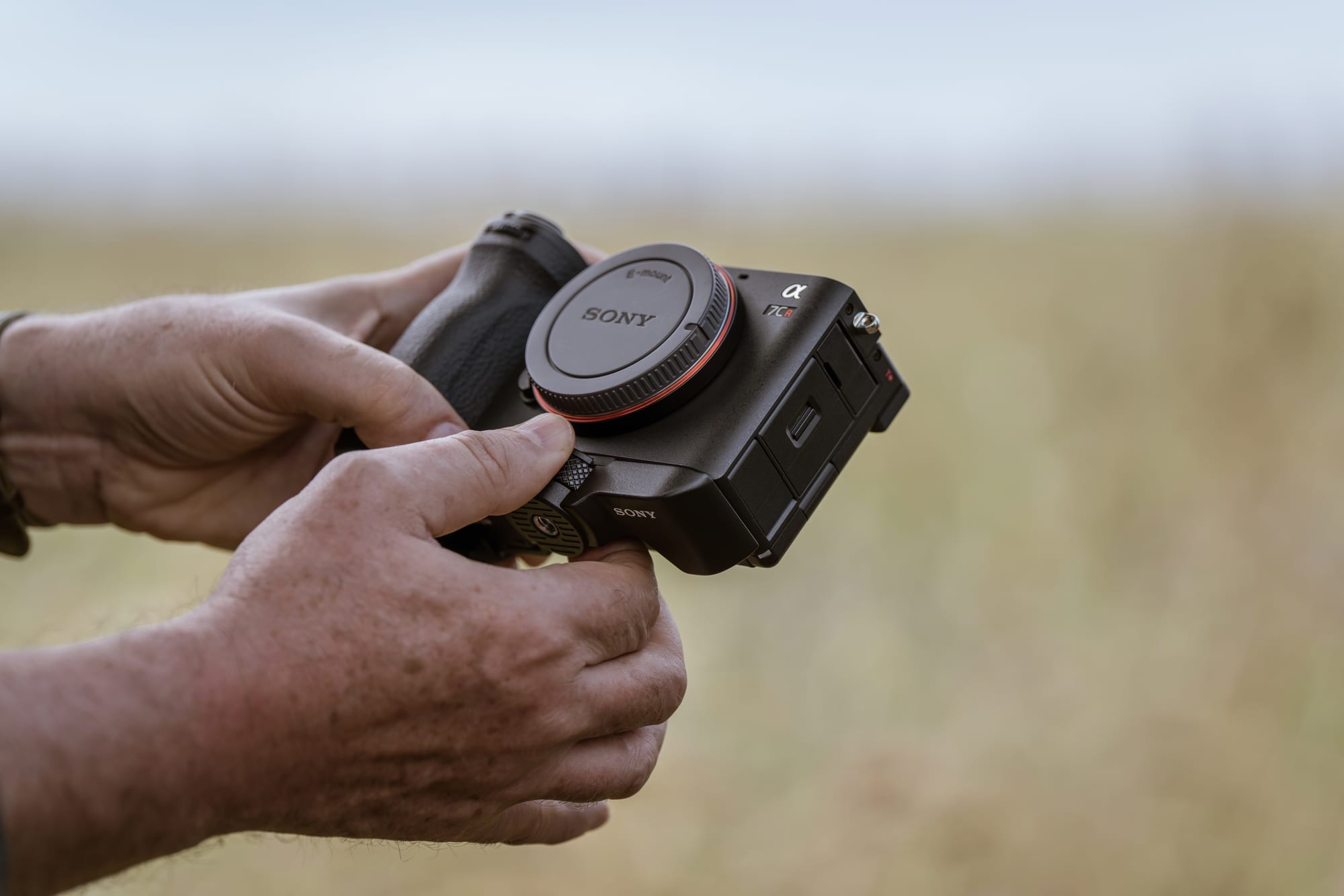 A full-frame camera body held in two hands outdoors, body cap on and no lens fitted &mdash; just the body.