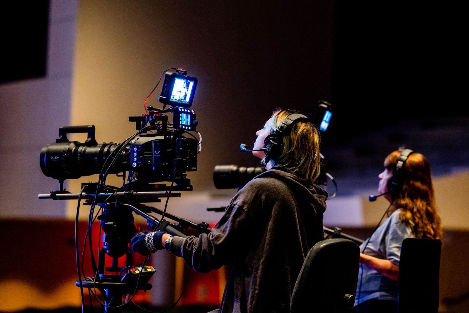 Two crew working a multi-camera shoot in a venue, both wearing intercom headsets —
                                  one operating a camera rig, the other seated at a second position beside them.