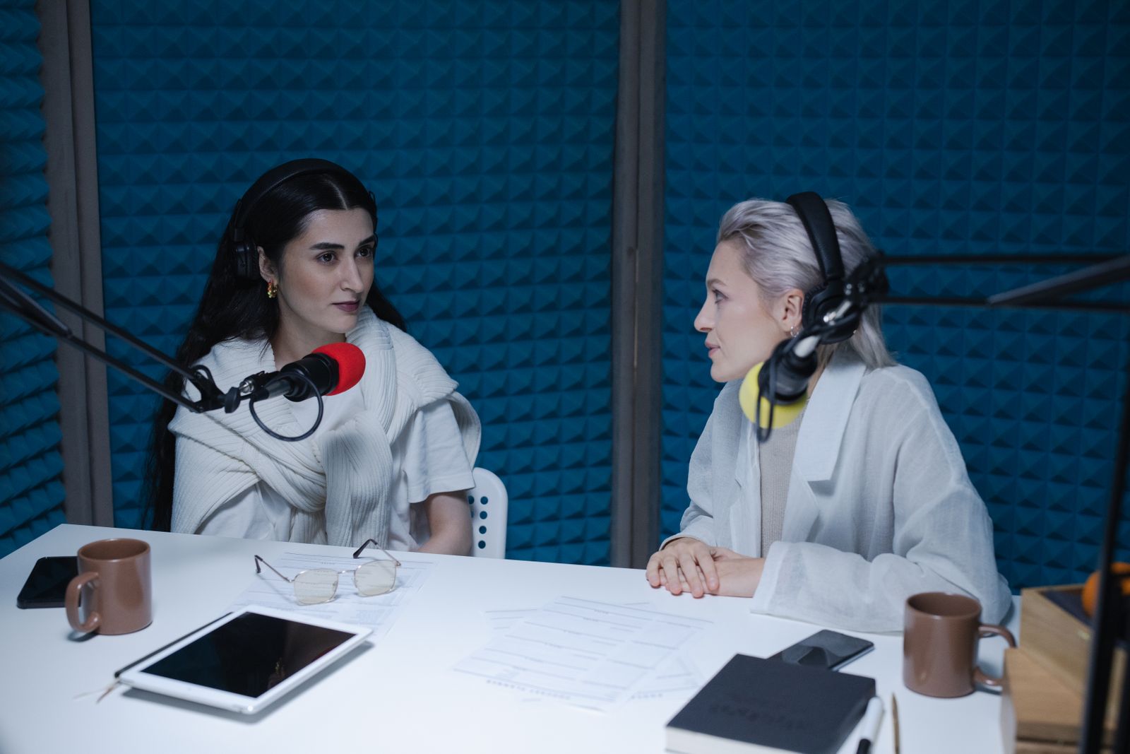 Two people seated across a table, each with their own microphone and headphones, in a treated room.