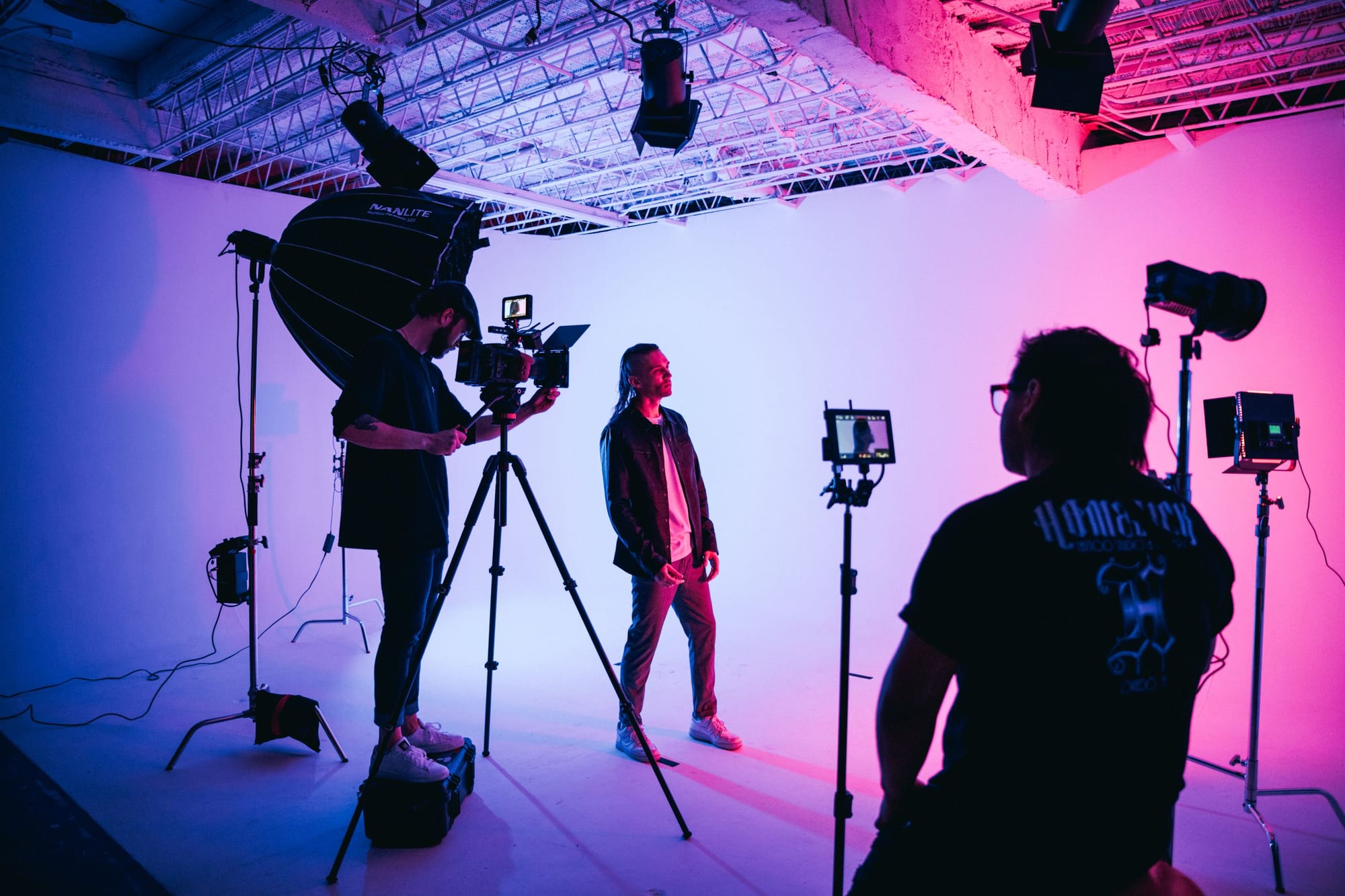 A studio shoot: an operator with a monitor-rigged camera on a tripod, a subject on a lit cyc, with crew and lights around them.