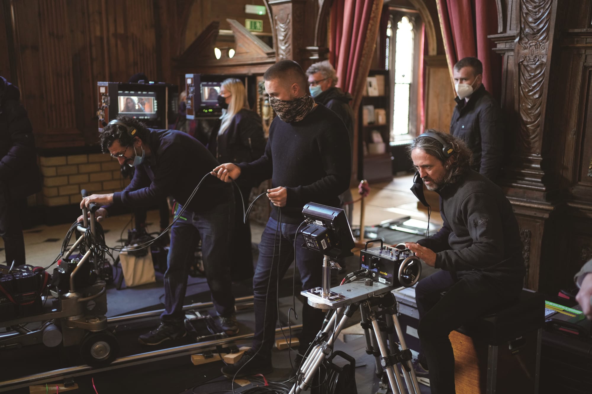 A film crew around a built-out cinema camera on a dolly and remote head, on a controlled set.
