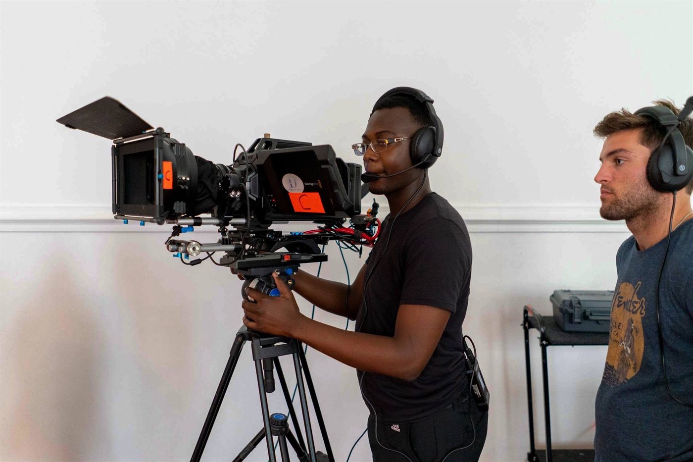 A camera operator on a cine rig and a second crew member beside him, both wearing intercom headsets.