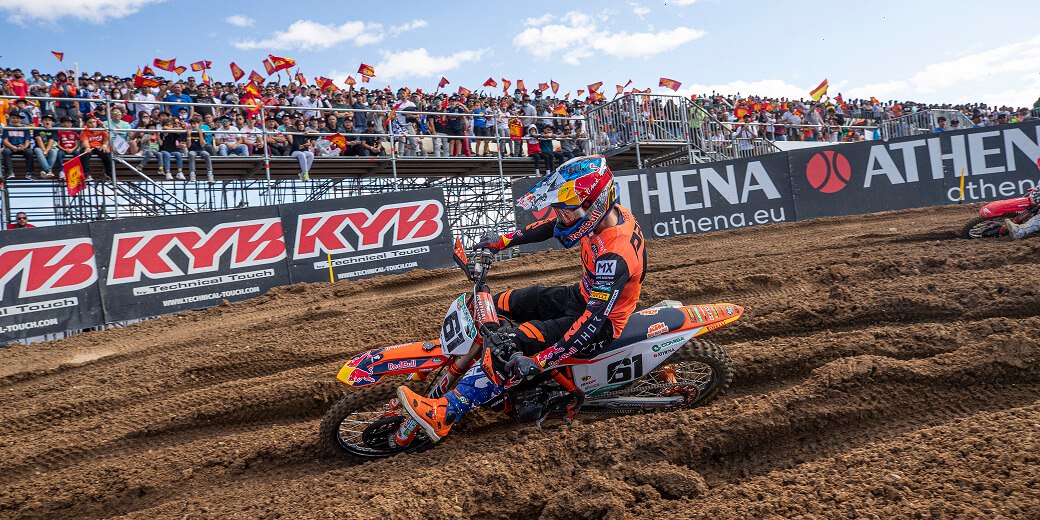 A motocross rider cornering in front of a packed grandstand at a live race meeting.