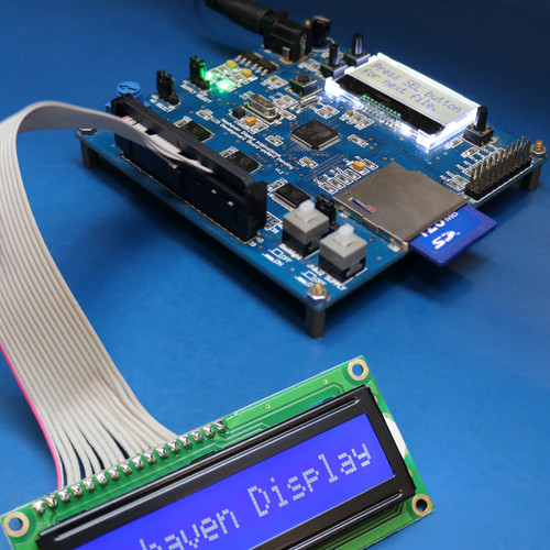 The blue development board is connected to an LCD with a dark-blue screen and white text via a gray ribbon cable.