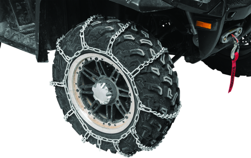 A high-resolution studio product photograph of QuadBoss Small V-Bar Tire Chains mounted tightly around an aggressive black ATV/UTV tread tire, set against a clean white background. Displayed at a front-three-quarter angle, the heavy-duty galvanized steel chain harness wraps securely across the tire's outer tread and sidewall, highlighting the sharp V-bar studs designed for maximum ice and snow traction. The blue plastic QuadBoss storage case sits neatly to the left of the mounted tire assembly.