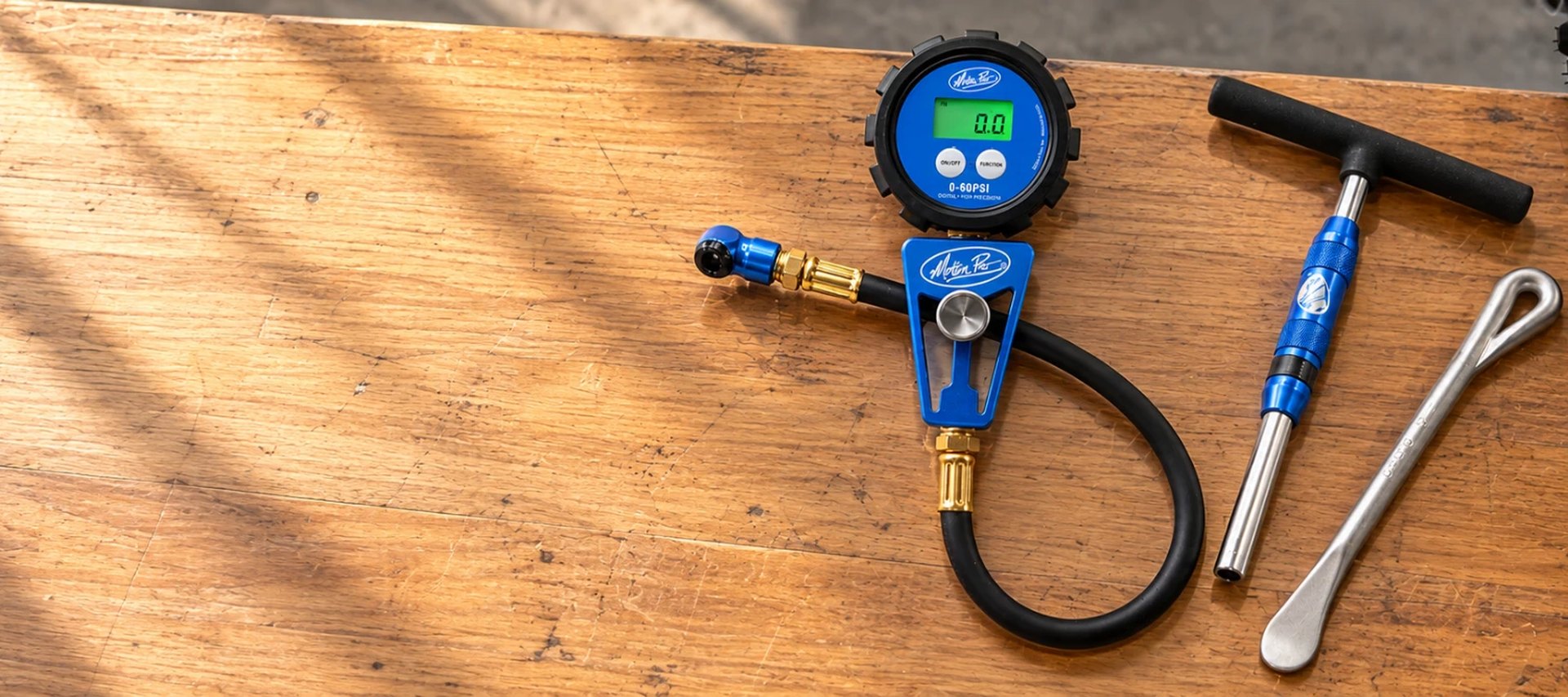 Motion Pro digital tire pressure gauge, T-handle wrench and tire spoon on a workbench next to a dirt bike wheel