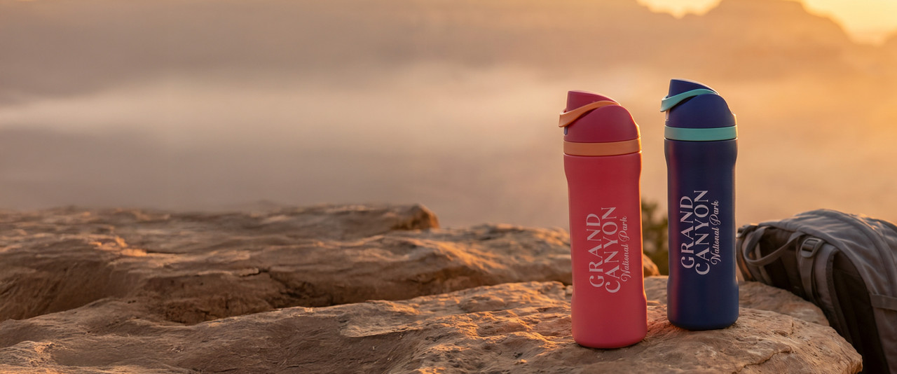 Pink and blue Grand Canyon water bottles sitting on the rim of the canyon with a backpack beside them.