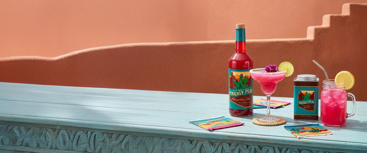 Prickly pear margarita and lemonade sitting on a turquoise table with an adobe building style background.