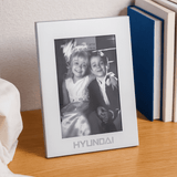A silver aluminium photo frame displaying a black-and-white photo of two children. The frame features a logo at the bottom.