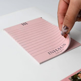 A pink notepad with lined pages is being peeled off a white magnetic whiteboard, which features a branding logo. A pink notepad with lined pages is being peeled off a white magnetic whiteboard, which features a branding logo.