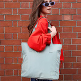 A woman with long hair holds a light blue tote bag against a brick wall, wearing a red jacket and sunglasses.