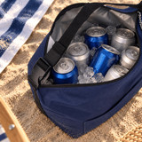 A navy cooler bag filled with ice and assorted cans, placed on sandy ground alongside a beach towel.