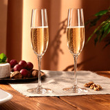 Two elegant, clear champagne flutes filled with sparkling wine, positioned on a wooden table with grapes and nuts.