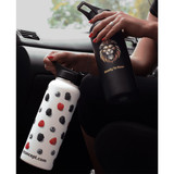 A drink bottle with a white body featuring a fruit design, alongside a black bottle with a logo. Both are in a vehicle.
