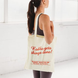A calico tote bag in cream colour featuring a logo and text. A person holds it while wearing activewear.