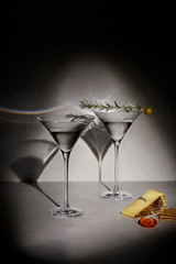 A set of two elegant clear martini glasses with a garnish, accompanied by cheese and crackers on a grey surface.