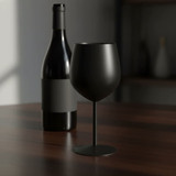 A black stainless steel wine glass stands beside a wine bottle on a wooden table. The setting has soft, ambient lighting.