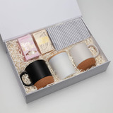A gift box containing two mugs (black and white), marshmallows, and a striped napkin on shredded paper filling.