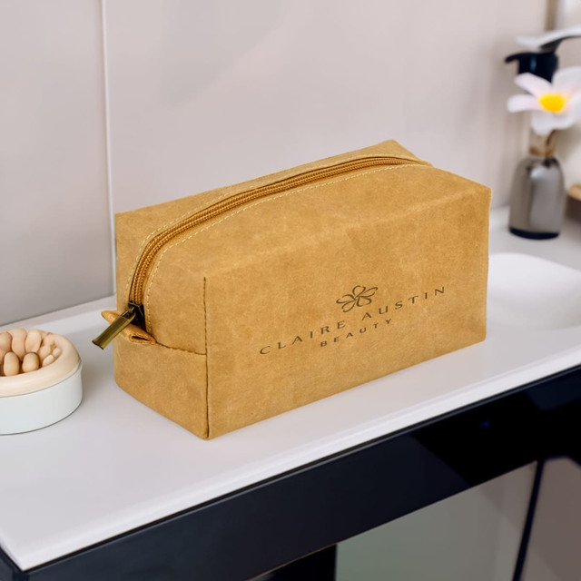 A tan kraft paper cosmetic bag with a zipper, featuring a logo, displayed on a bathroom countertop. A tan kraft paper cosmetic bag with a zipper, featuring a logo, displayed on a bathroom countertop.