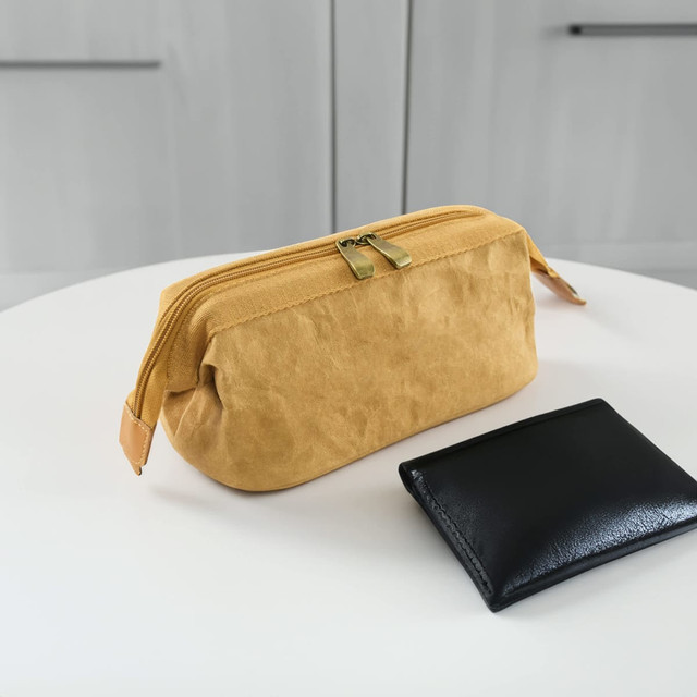 A yellow kraft paper cosmetic bag with a zip closure sits beside a black wallet on a white table. The bag has a logo.