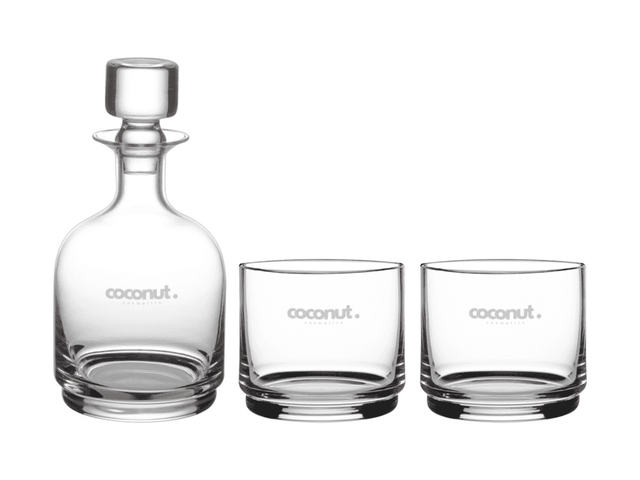 A glass decanter and two matching tumblers, all clear with a subtle logo.