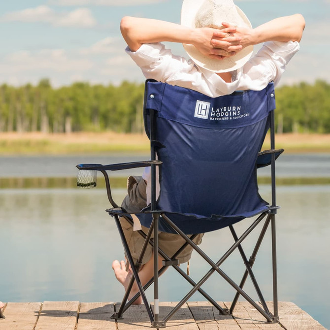 A blue folding chair with armrests, featuring a logo, set against a lakeside backdrop.