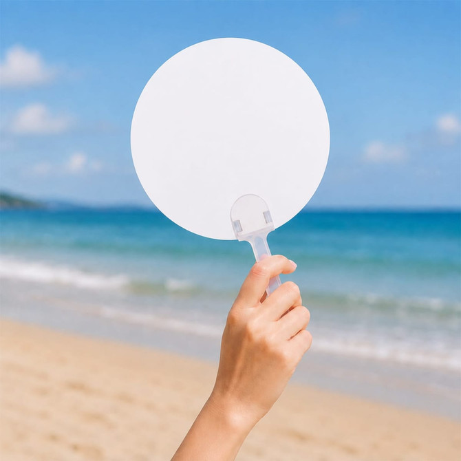 A handheld circular fan with a transparent handle against a beach backdrop and ocean waves.