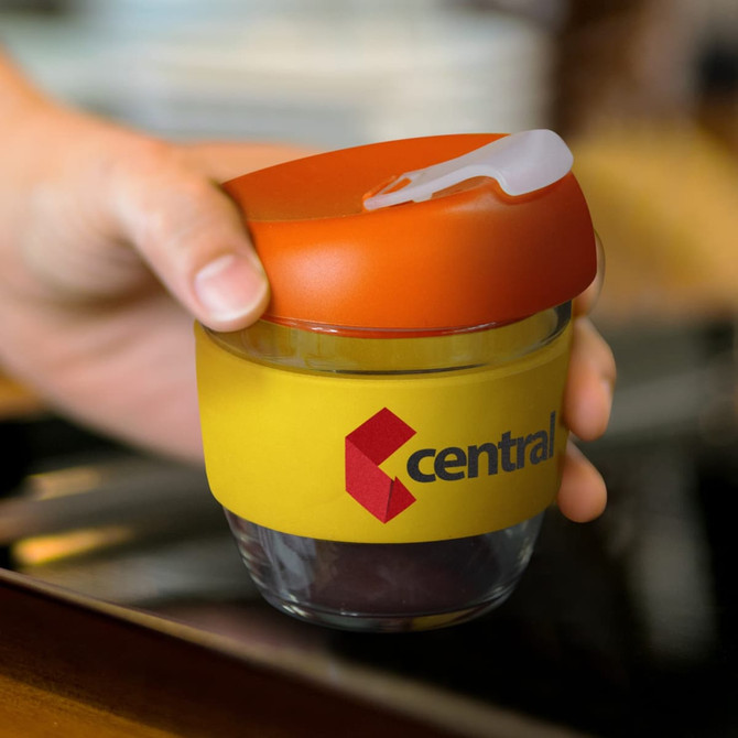 A drink bottle with a clear borosilicate body, orange lid, and yellow sleeve, featuring a logo.