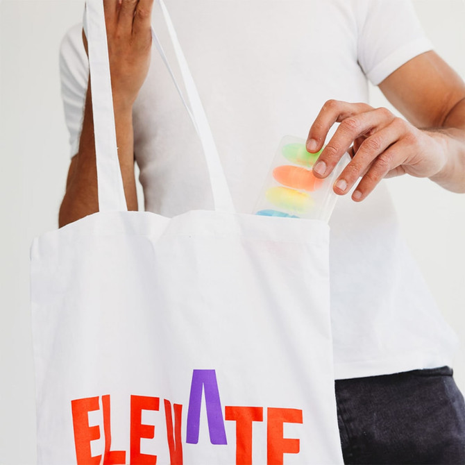 A white tote bag with colourful lettering, being held by a person as they retrieve a clear container with coloured items.