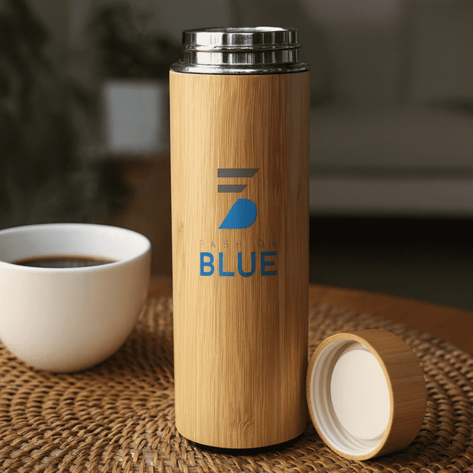 A drink bottle in bamboo finish with a silver lid and a logo. It sits on a woven surface near a cup.