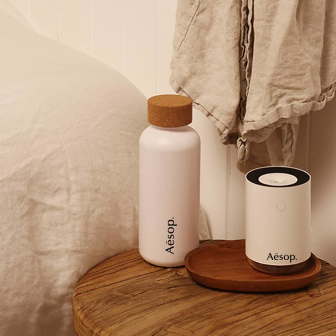 A drink bottle in a light pink colour with a cork lid, alongside a diffuser on a wooden surface.