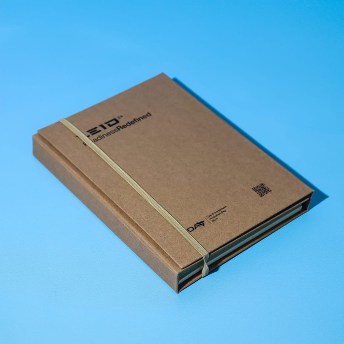 A brown A5 notepad secured with a rubber band, featuring a logo on the cover against a blue background.