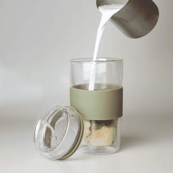 A glass coffee cup with a green silicone sleeve, being filled with milk from a metal jug.